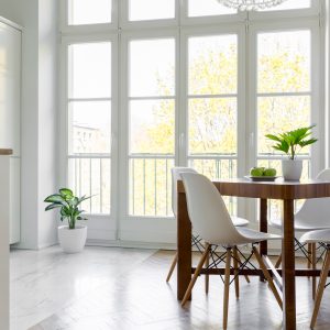 White chairs at wooden table with plant in bright dining room interior with window. Real photo